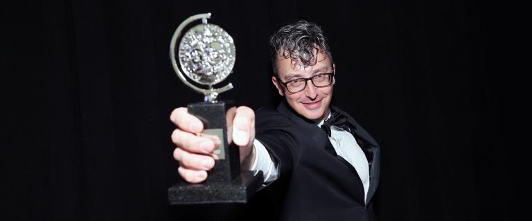 Beowulf Boritt dramatically lit wearing glasses and a tuxedo thrusts a trophy forward and up.