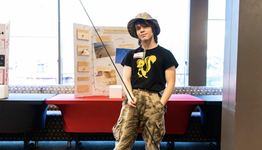 A person with long brown hair stands in front of a wall of large windows. The person is wearing a brimmed hat, camouflage pants, and a black T-shirt with a yellow cartoon drawing of a skunk on it. The person is holding a long fishing pole.