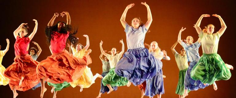 A large group of people dancing on a stage. They are all wearing different color dresses while jumping in the air with their hands over their heads in unison.