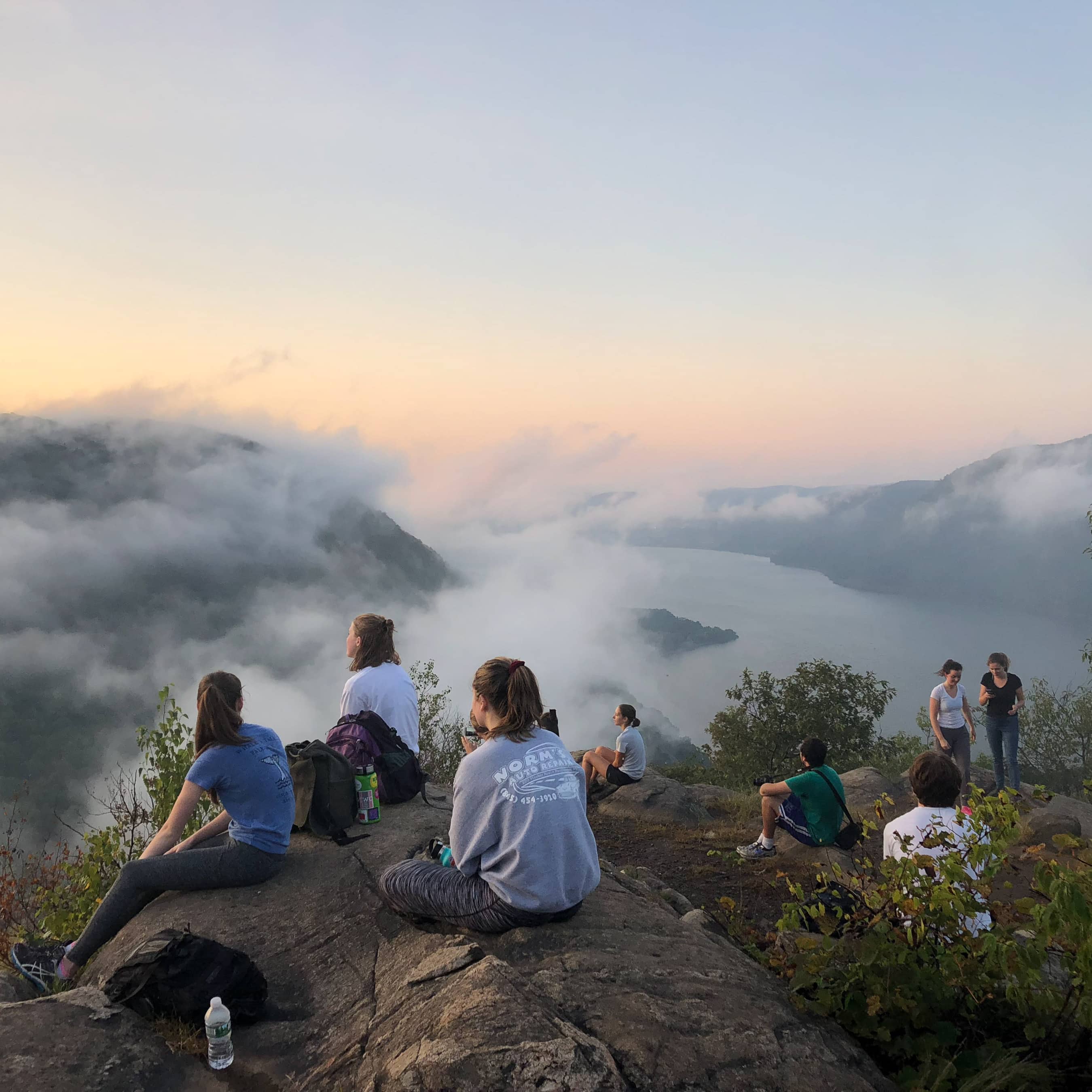 A group of students sit on a rocky open area on top of a mountain, overlooking the fog-covered Hudson River.