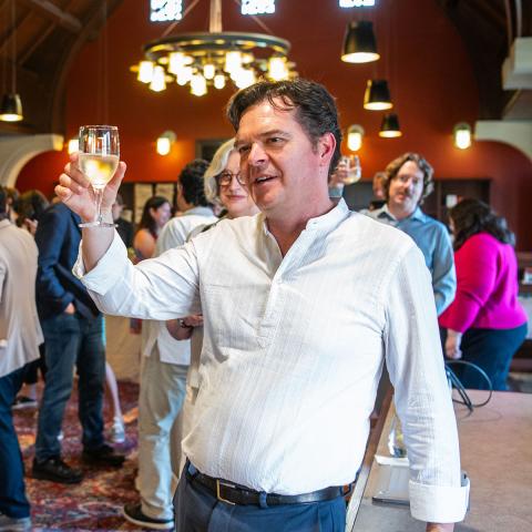 Man standing holding up a glass to toast in a large room with people socializing and eating. 
