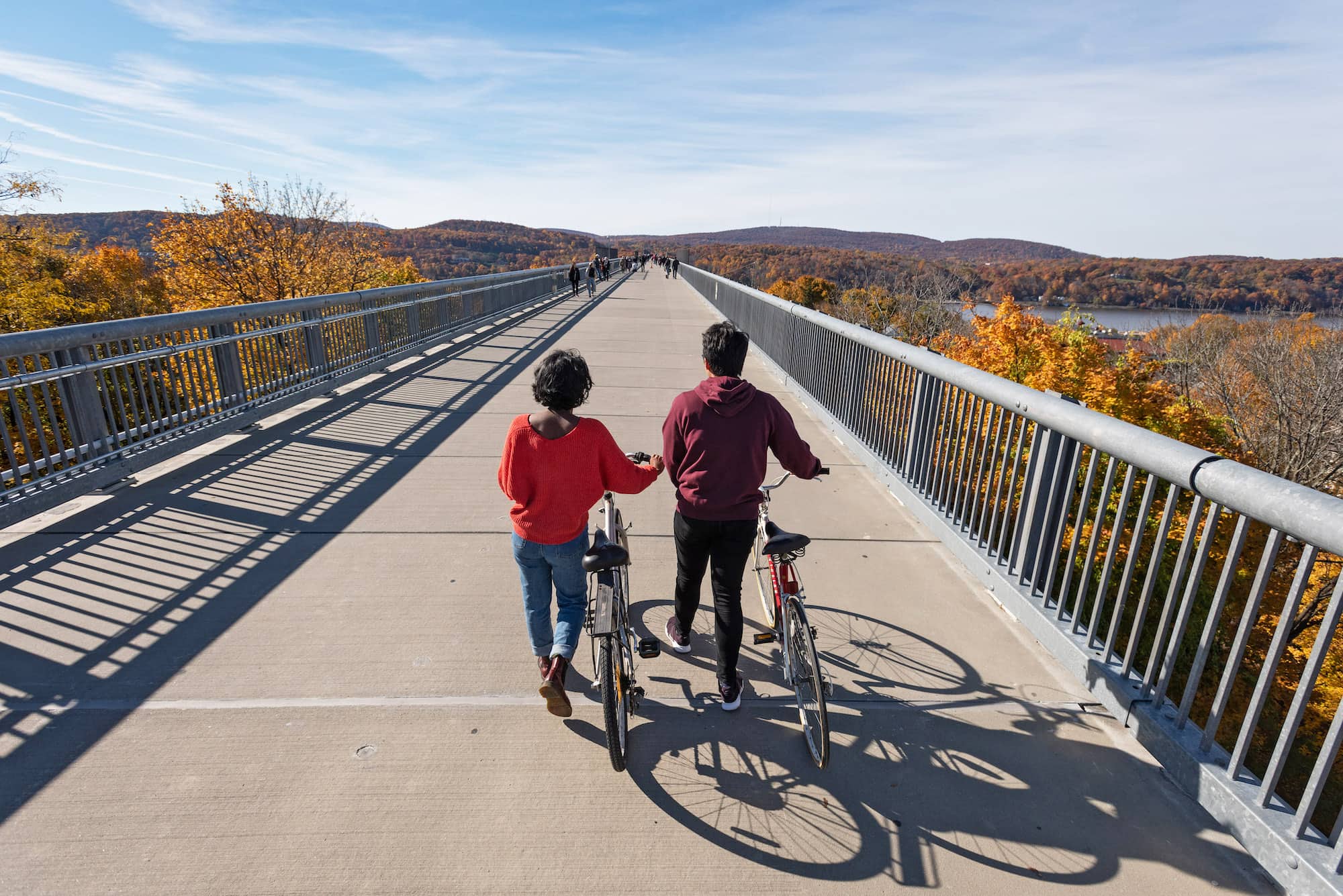 Two students facing away from the viewer wheel their bikes along a long concrete pathway on a bridge overlooking the Hudson river.