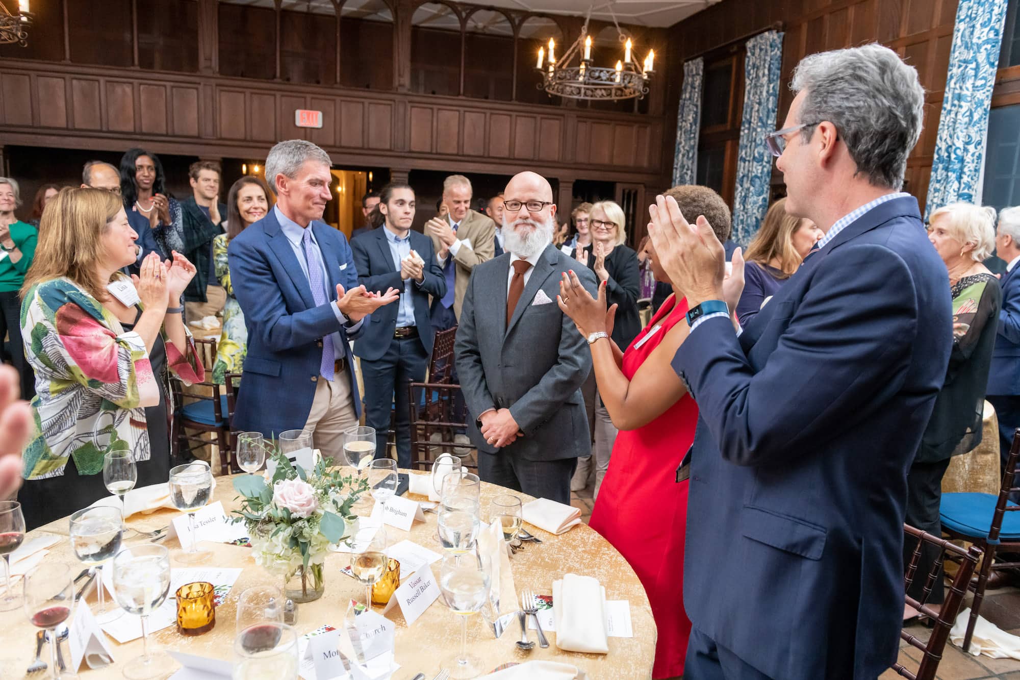 A group of people stand in a wood-paneled dining room applauding Professor Robert Brigham, a person with a bald head and a full white beard and mustache, wearing a suit.