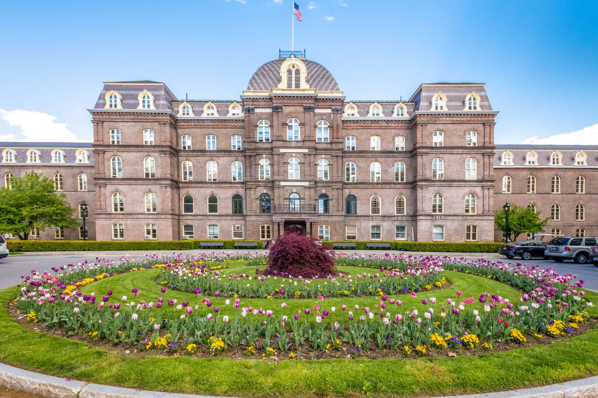 The front of Main Building on the Vassar campus, a large red brick building. In front of the building lies a circular garden area adorned with various types of colorful flowers.