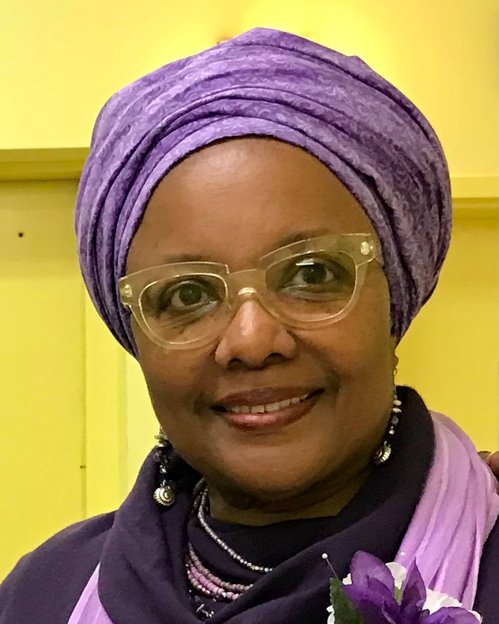 A person wearing a purple head covering, glasses, and a purple scarf smiles at the viewer.