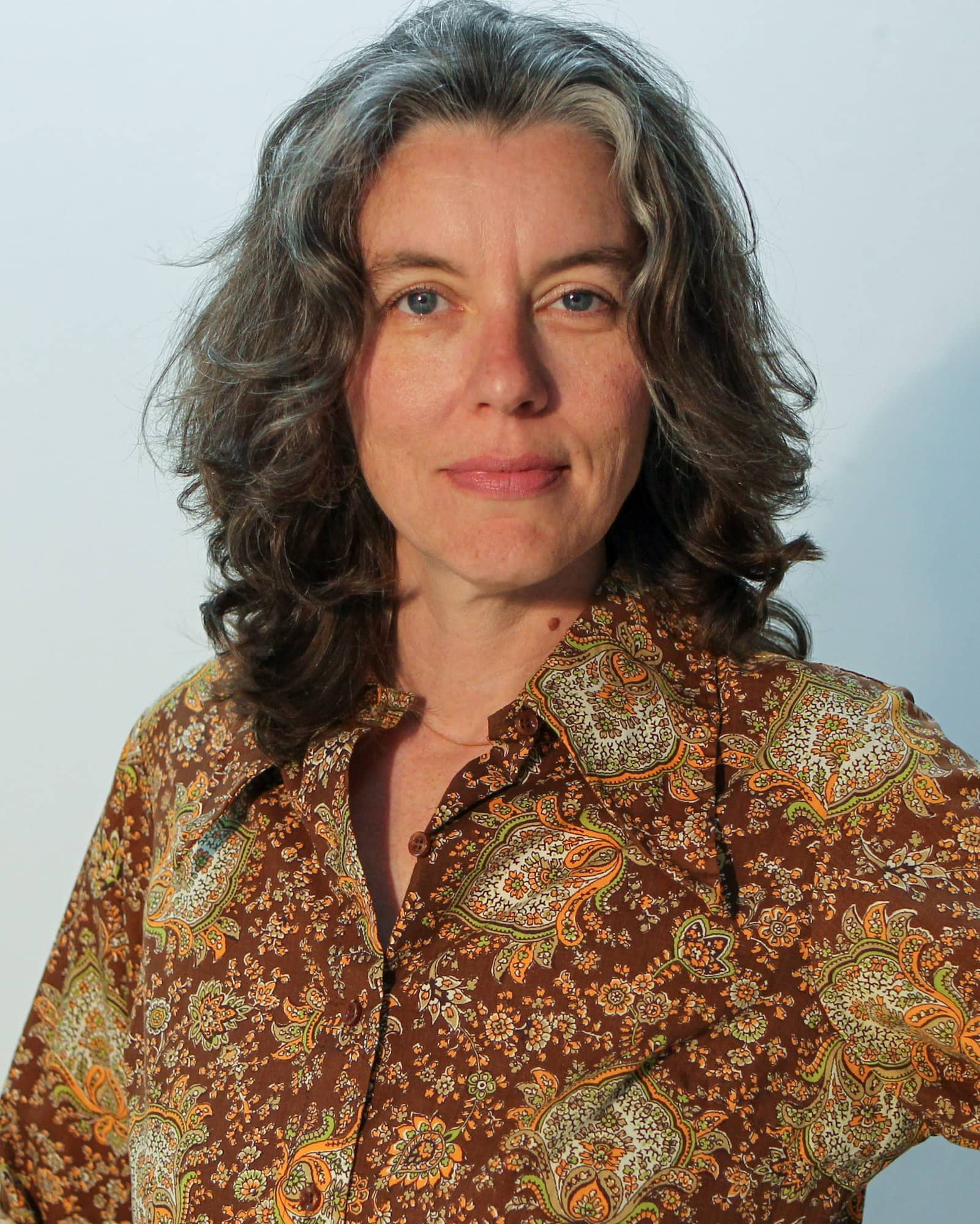 A person with shoulder length grey black wavy hair and a patterned collared shirt looks at the viewer.