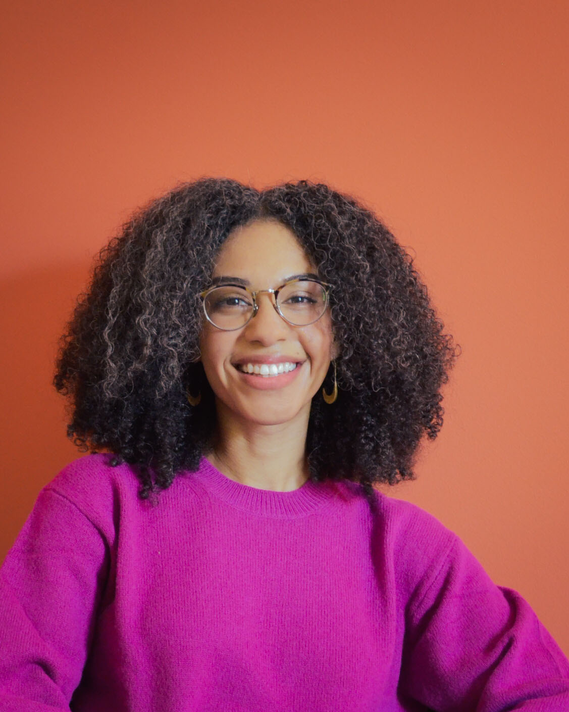 A person with curly shoulder-length hair wearing glasses and a pink sweater smiles at the viewer.