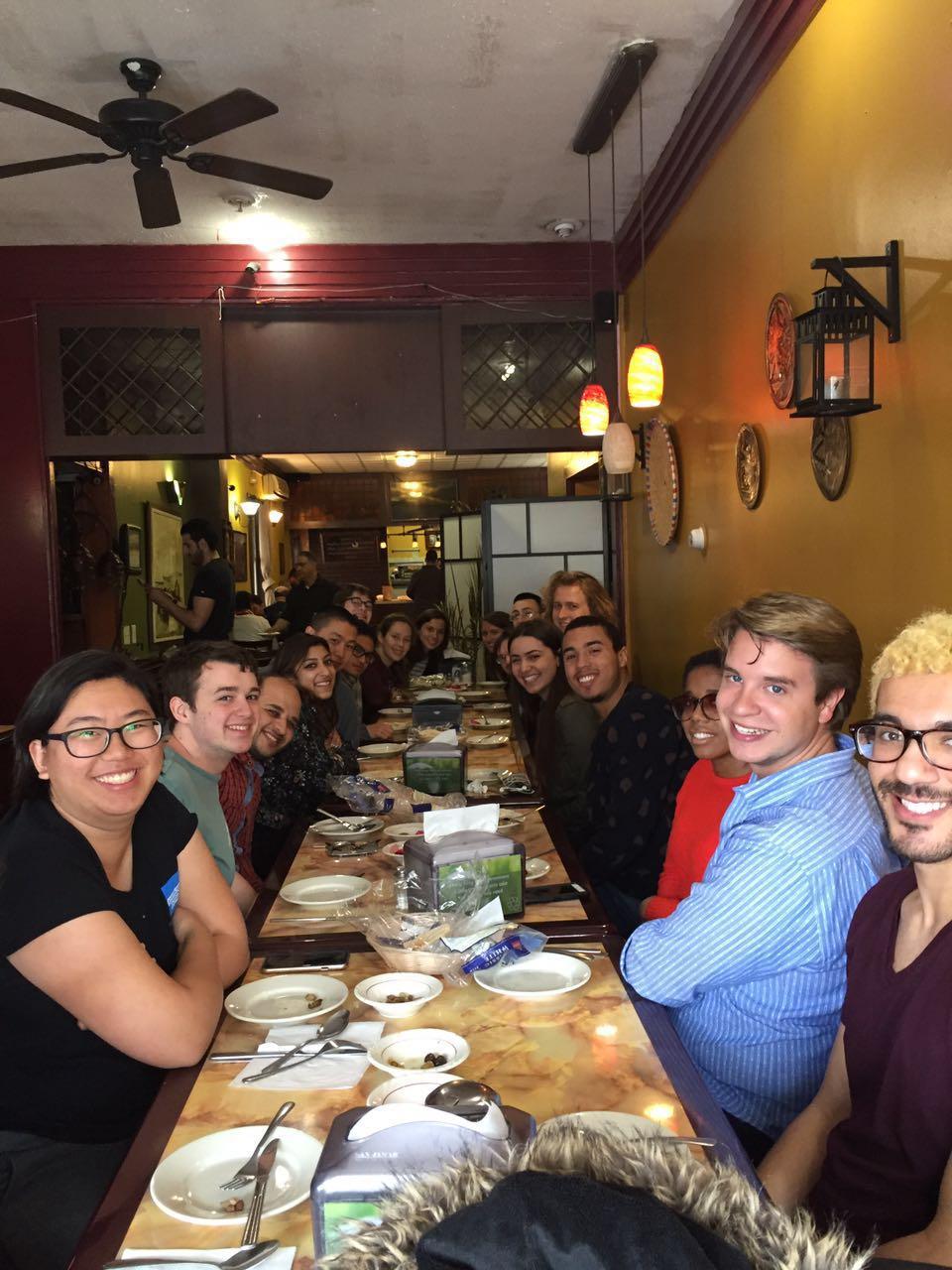 A large group of students and faculty sit at a table in a restaurant, smiling at the camera