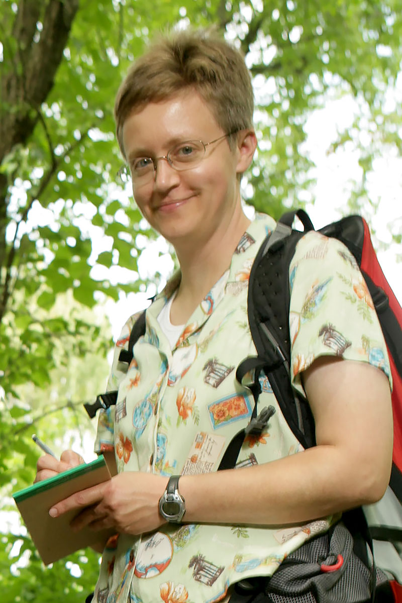 Kirsten M. Menking wearing a light patterned shirt and eyeglasses with a tree in the background.