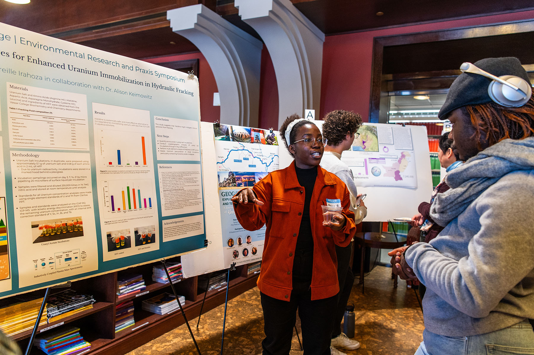 Person in a red sweater is pointing to a poster presentation on an easel while another person looks on.