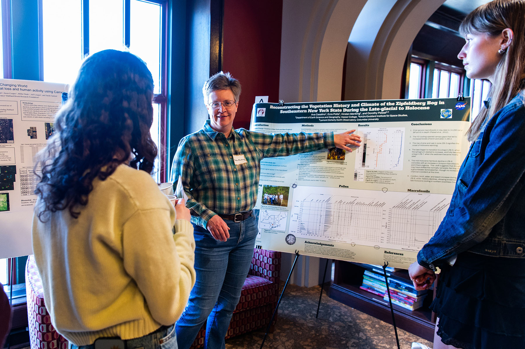 Person in a flannel shirt and jeans is pointing to a poster presentation on an easel while another person looks on.