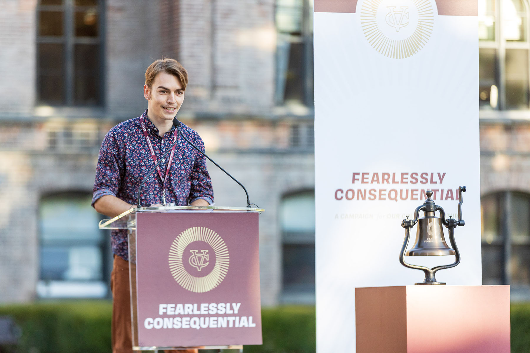 Person standing at a podium on a stage. A banner is draped over the podium that reads, "Fearlessly Consequential." In front of the podium is a silver bell mounted on a block of wood.