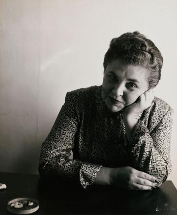 Vassar alum Elizabeth Bishop ’34, sitting with one arm on her cheek and one on a table and wearing a printed shirt, in black and white.