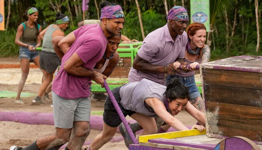 Five people dressed in a purple theme sweating. They are pushing a sled on a beach loaded with large wooden chests as if in a race. 