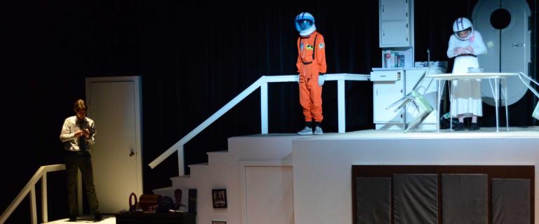stage with three actors, two wearing space helmets the other looking at a book 