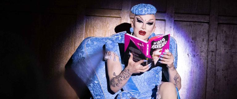 Sasha Velour seated with a book in the spotlight reading aloud.