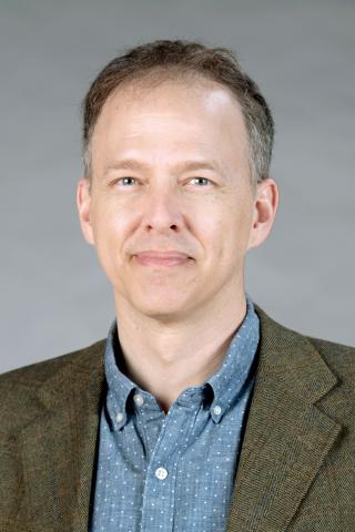 Mark Cleaveland wearing a light teal collared shirt and an olive colored jacket against a gray background.