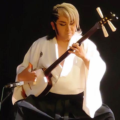 A seated performer wearing robes strums a stringed instrument.