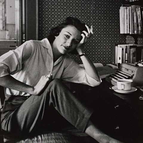 Rollie McKenna sitting with her left hand on her head, holding a cigarette, with books and a typewriter in the background, in black and white. 
