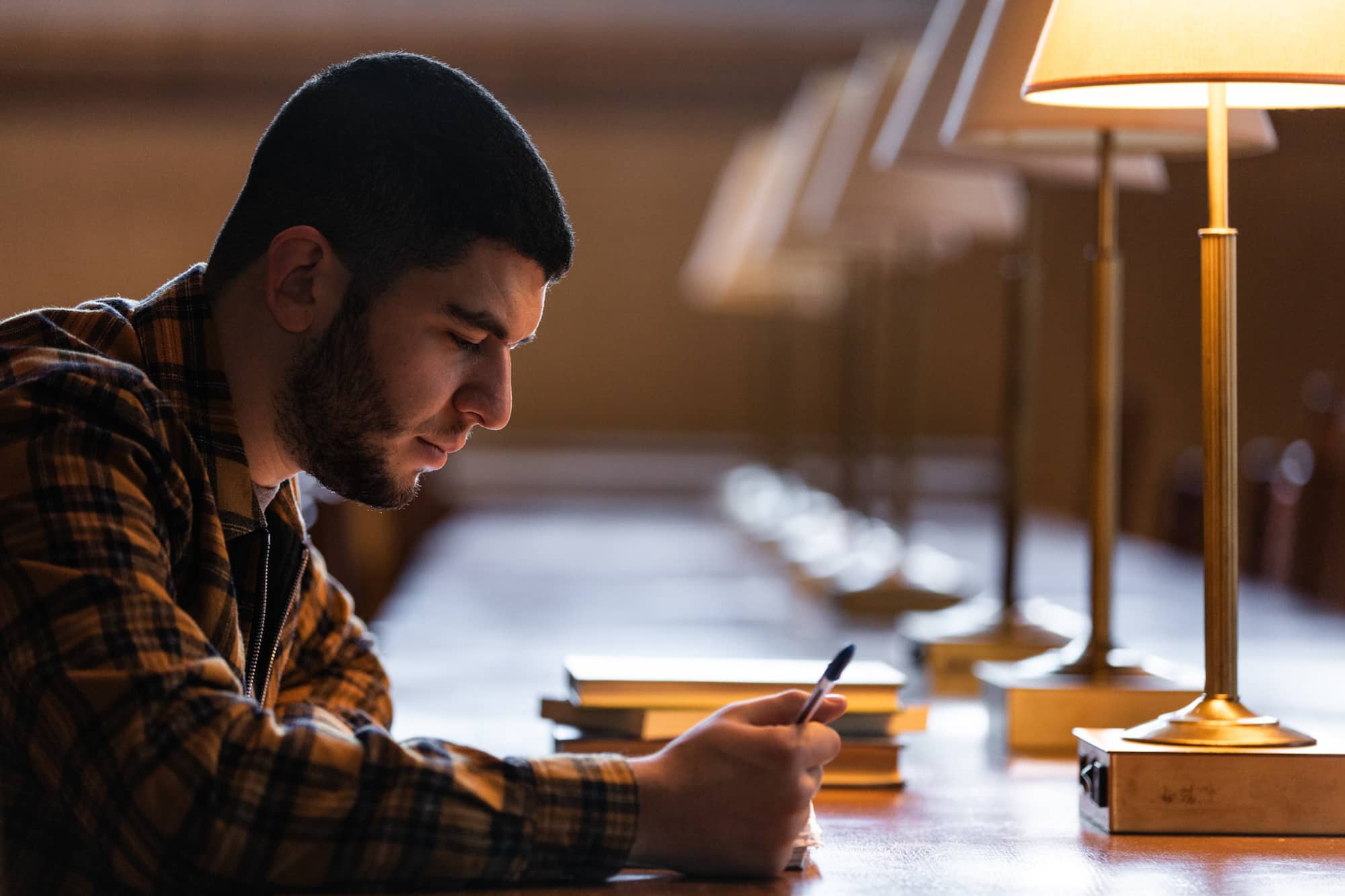 A student with dark hair and a beard sits at a long table, focusing on papers.