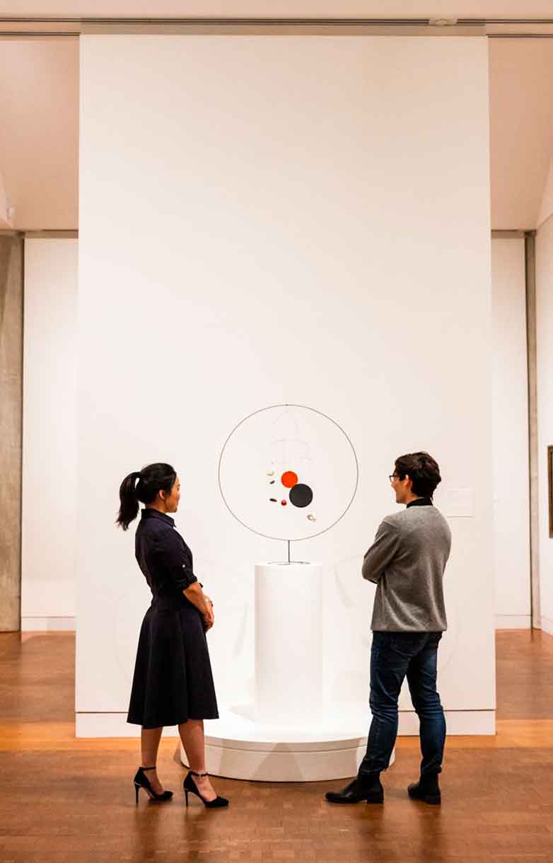 Two students stand in a large art gallery, looking at a small sculpture made of wire and colored metal.