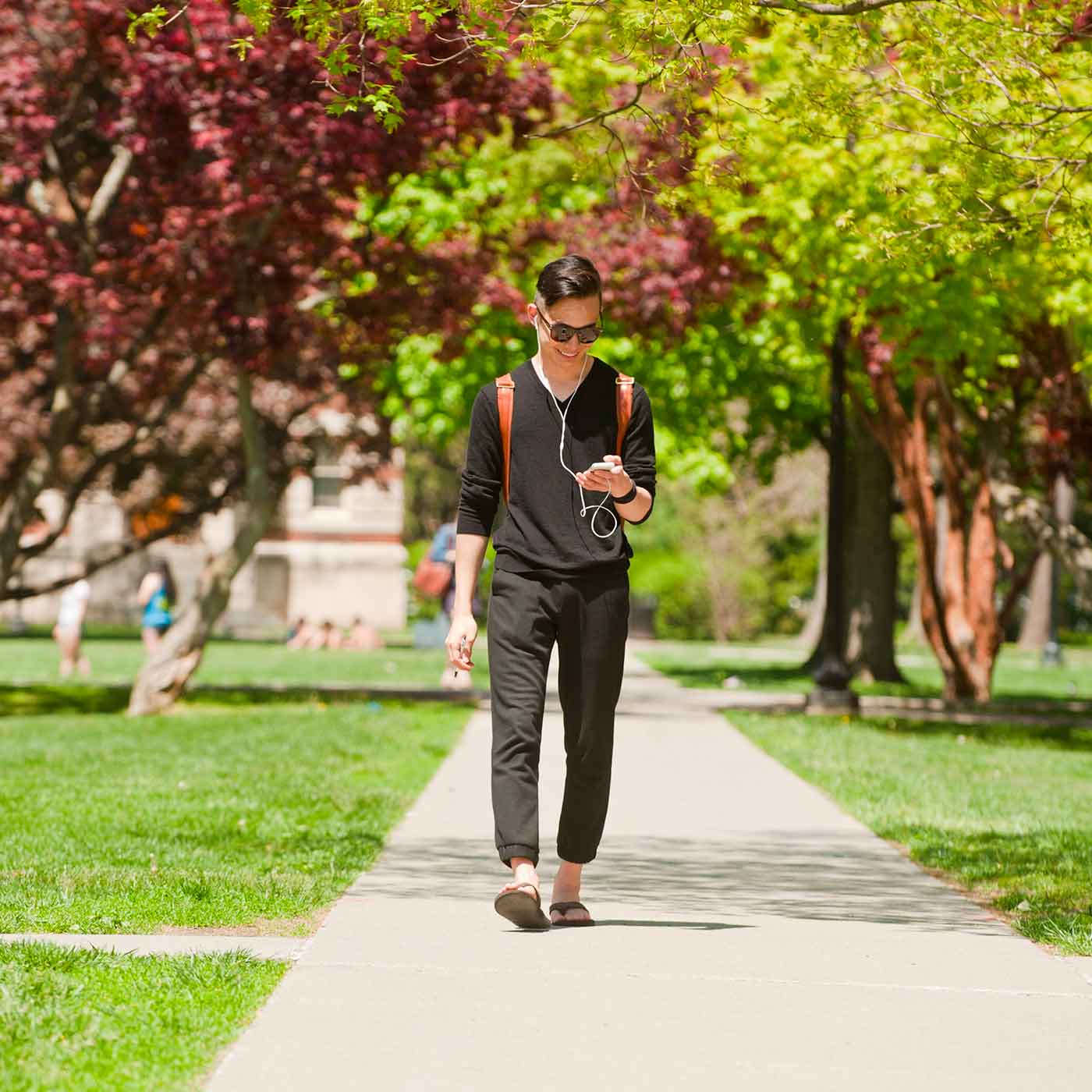 Student walking in the sun wearing earbuds and looking at phone.