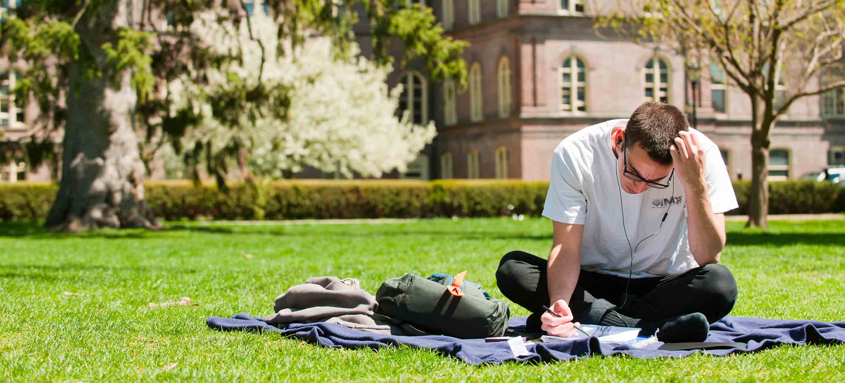 A student sits on a blanket in front of Main Building on a sunny day, looking down at some papers.