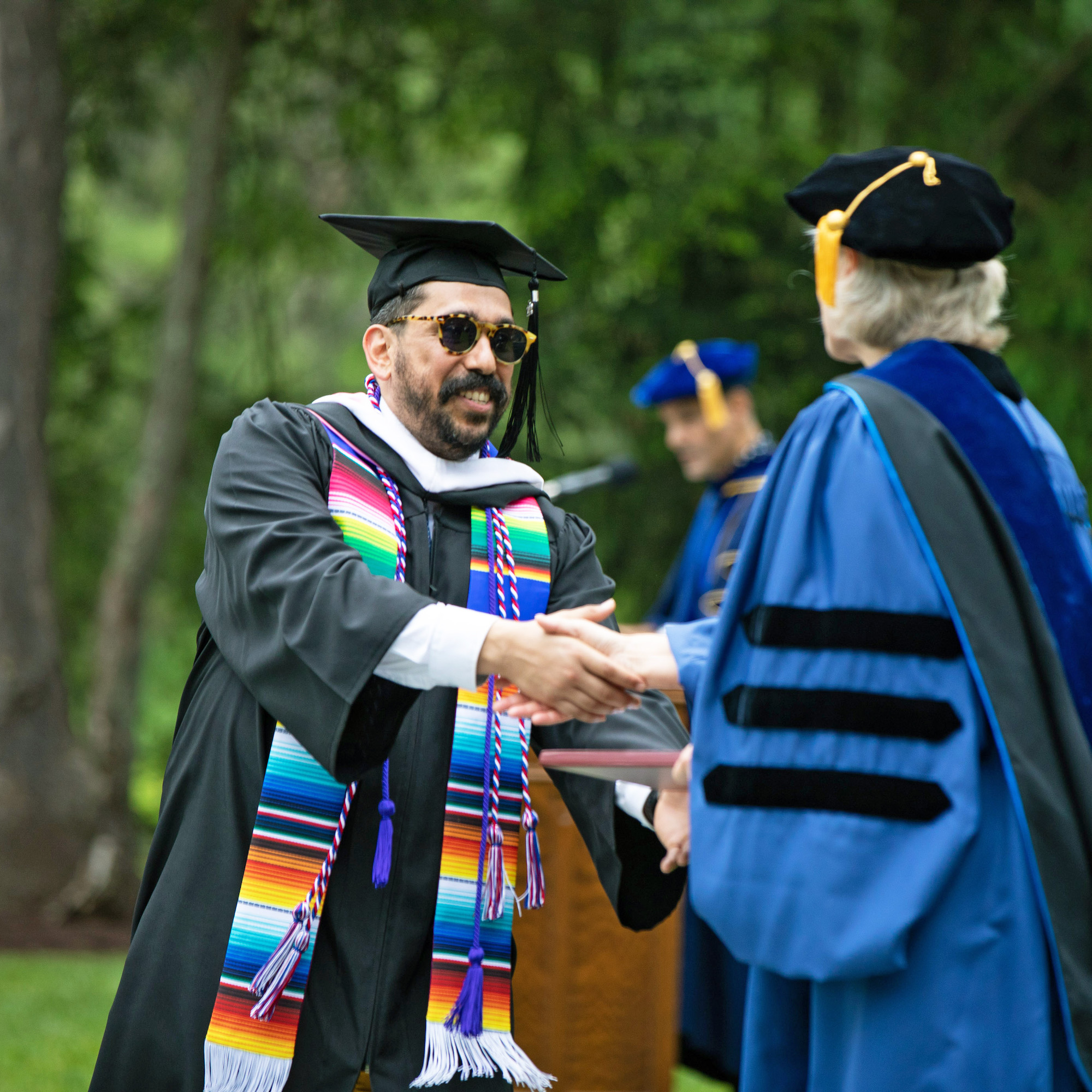 A graduate with sunglasses and dark facial hair wears a black robe with a bright, multicolored scarf, and shakes hand with President Bradley, who is wearing a blue robe and a black cap.