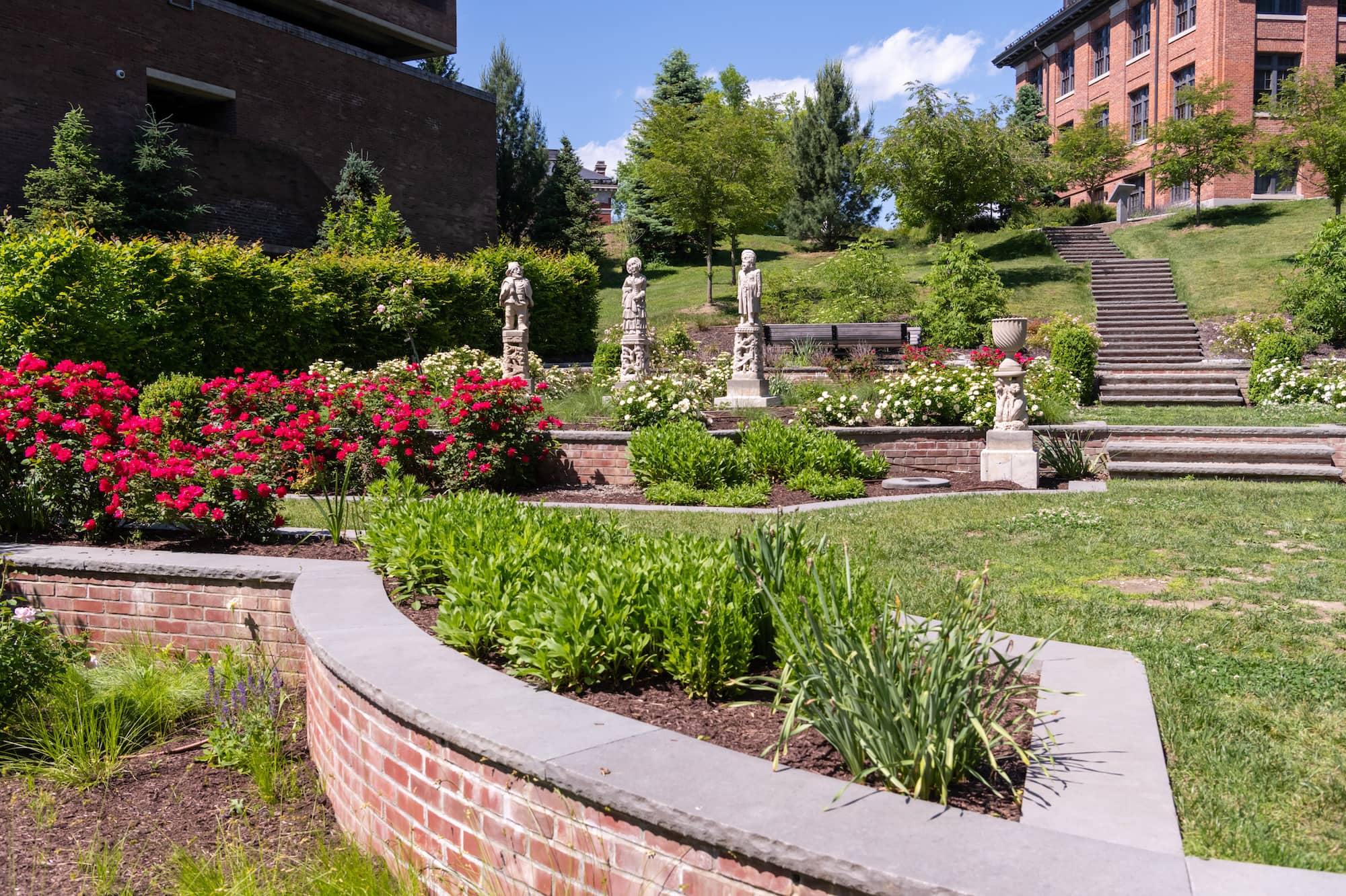 A large, open garden area with several stone statues and brick buildings in the background.