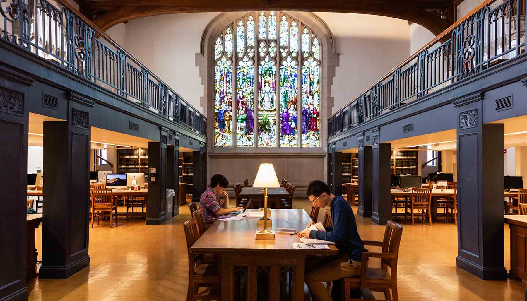 Students read in a spacious library reading room with a large stained-glass window behind them