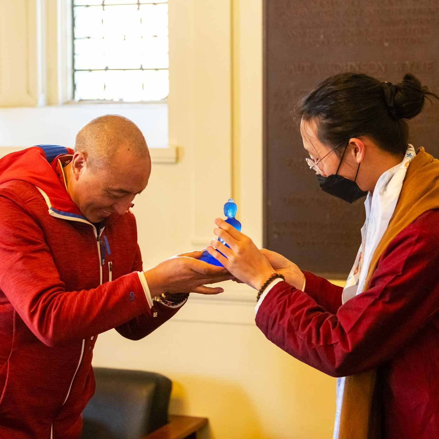 Two people wearing red robes exchange a small glass blue Buddha figurine