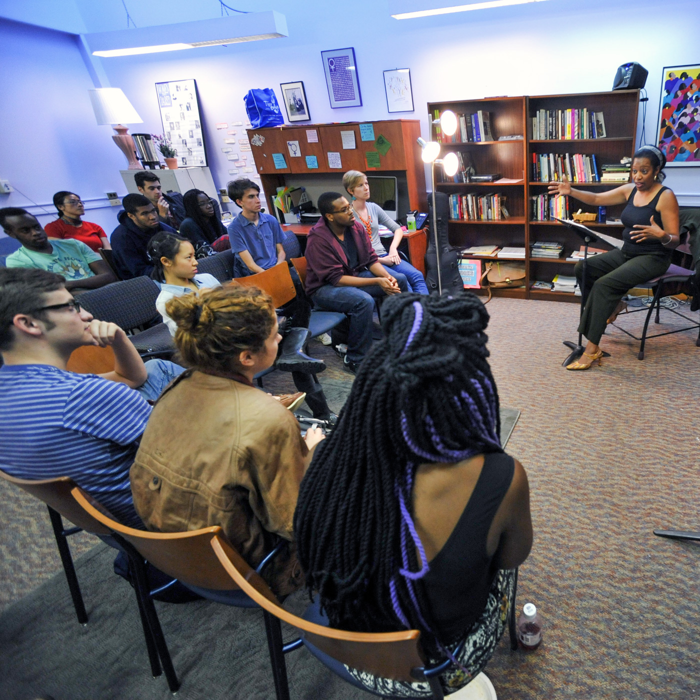 Group of 12 seated people sitting in front of and listening to a seated person speaking and gesticulating with their arms and hands