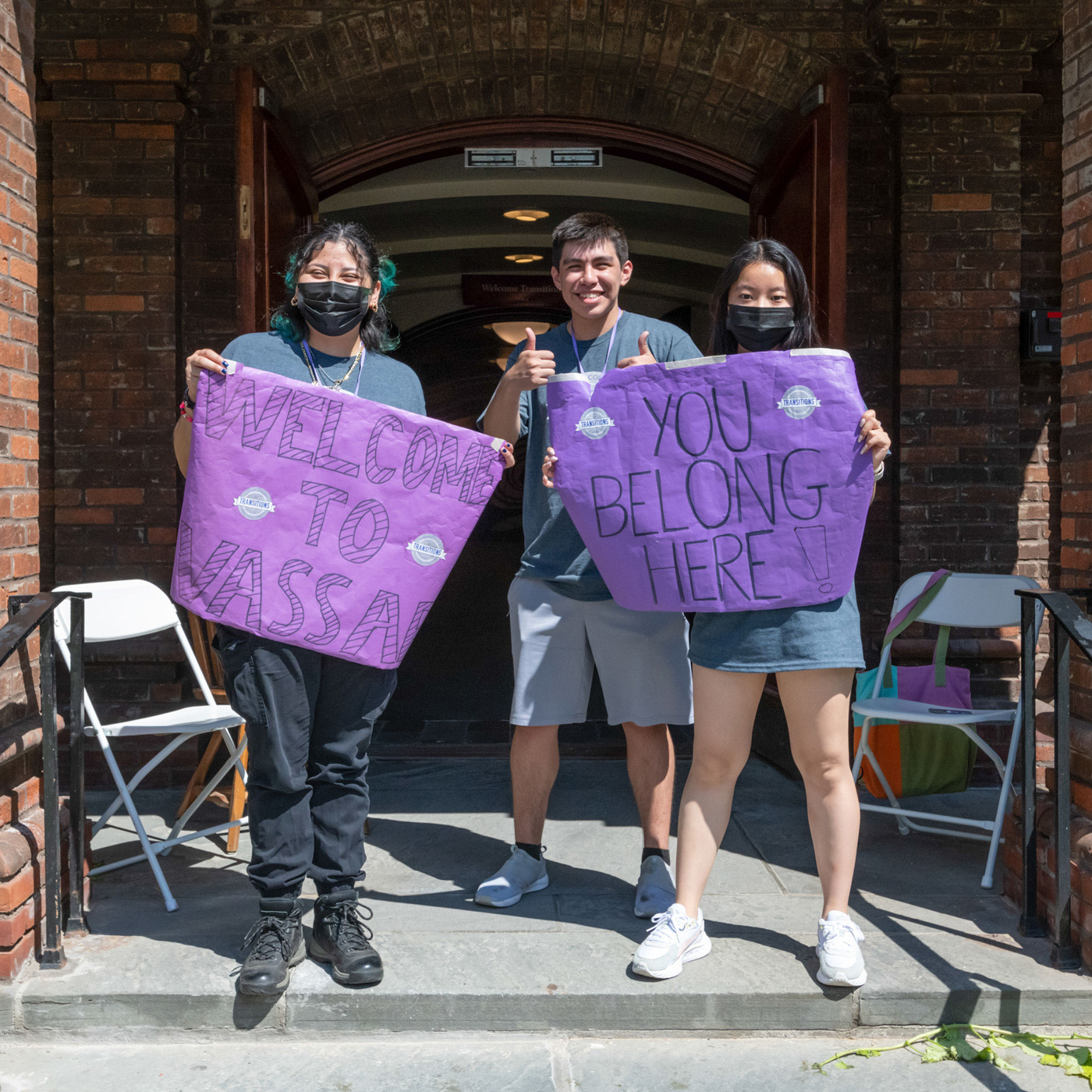 Three people at the entrance to Vassar College Main Building holding signs that read, Welcome to Vassar and You Belong Here. Two people are wearing face masks and one is not and smiling.