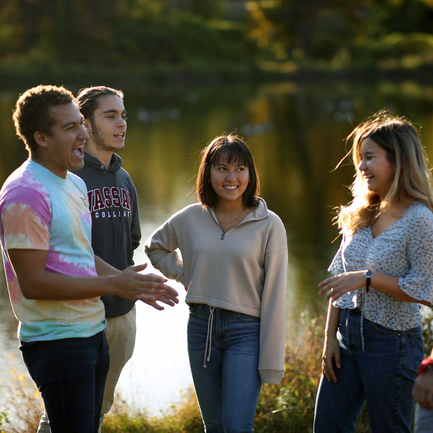 Evening sunlight shines on four people smiling and laughing in front of Sunset Lake on Vassar Campus.