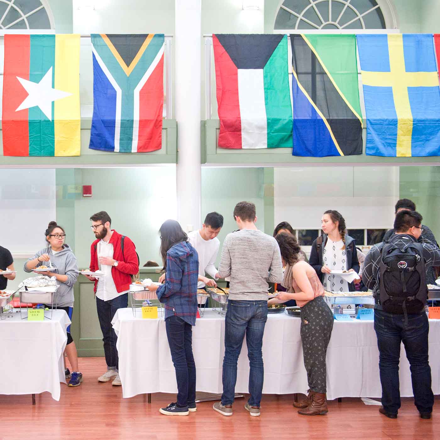 People stand in a line while food is served in a large room with flags of different countries