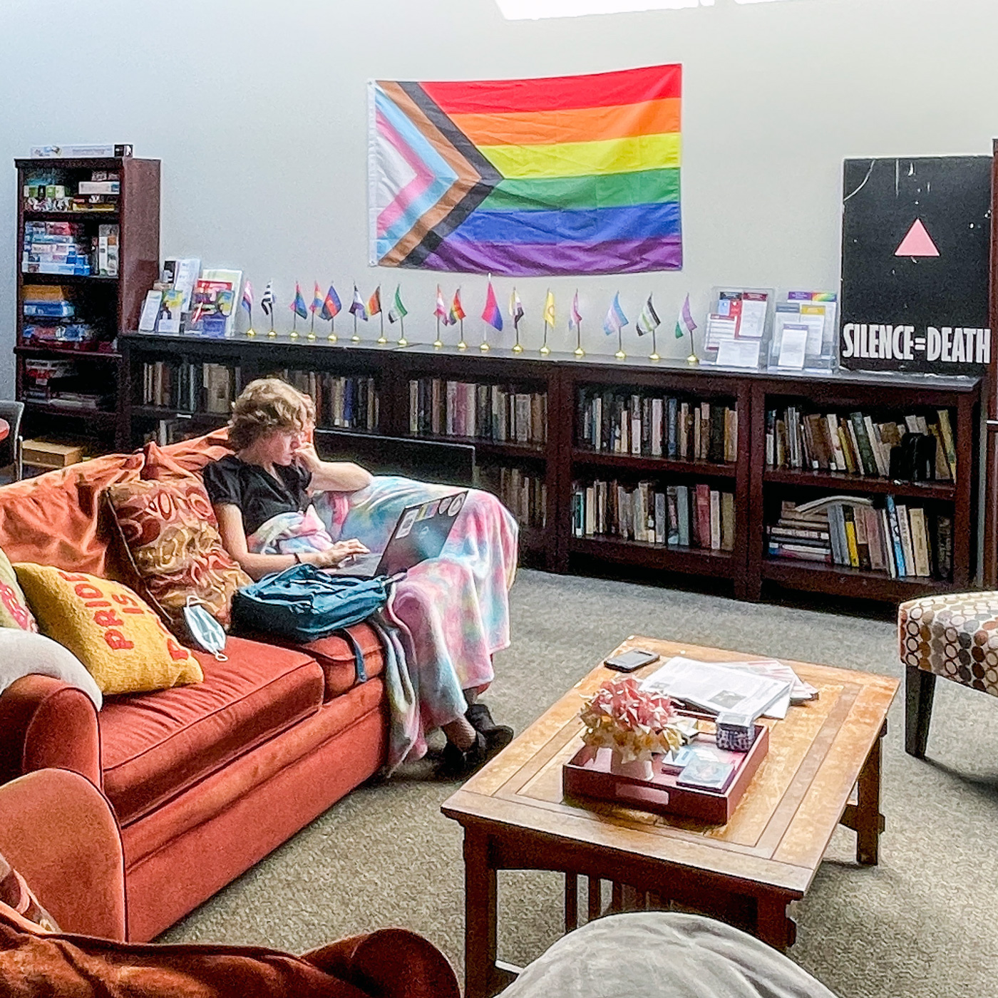 Person on a couch with laptop in a sunlit lounge with books, rainbow flags, posters, chairs and coffee tables.