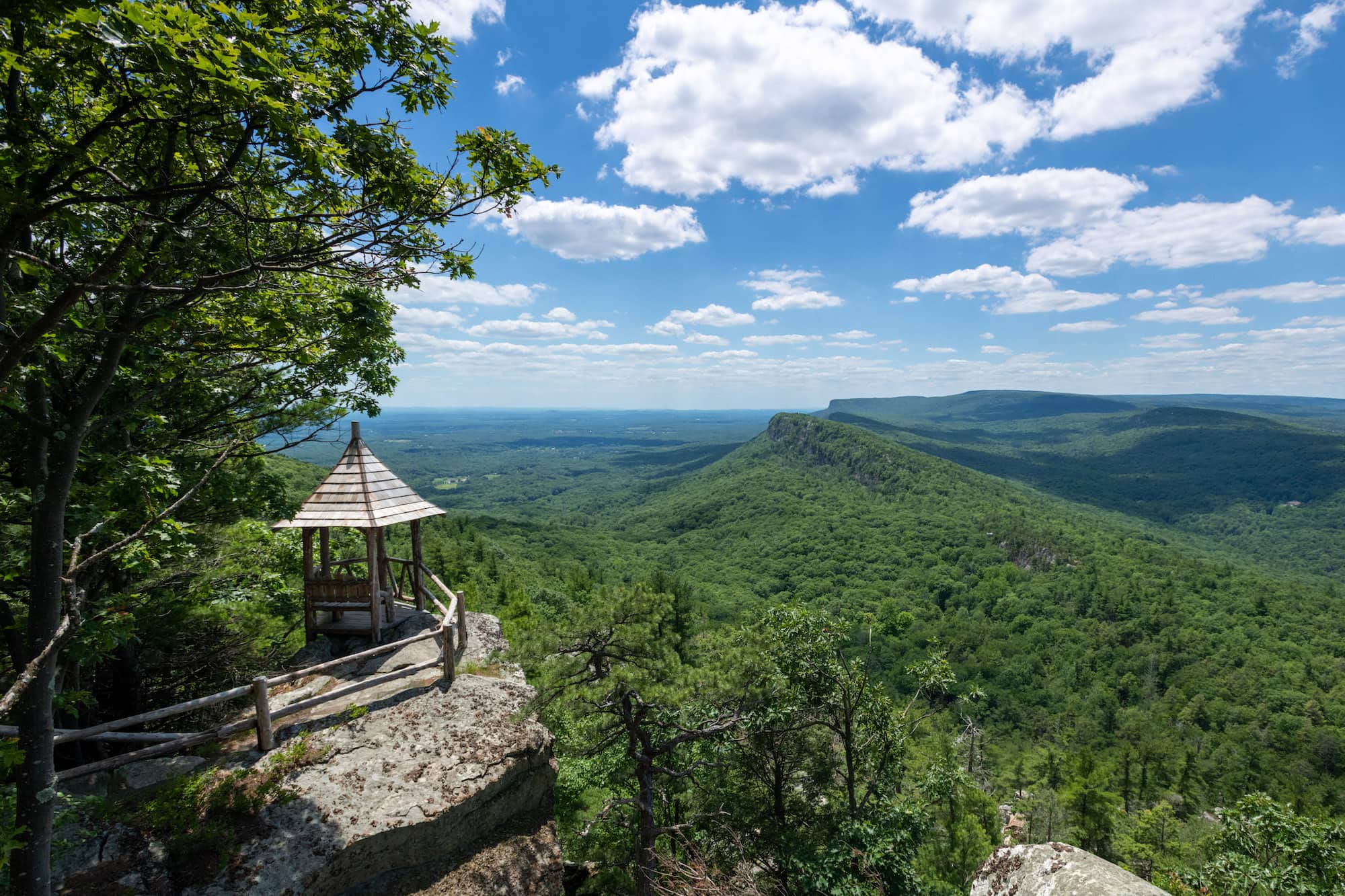 A view from a mountaintop out over a vast open space, with blue sky and forest-covered mountains below and in the distance.