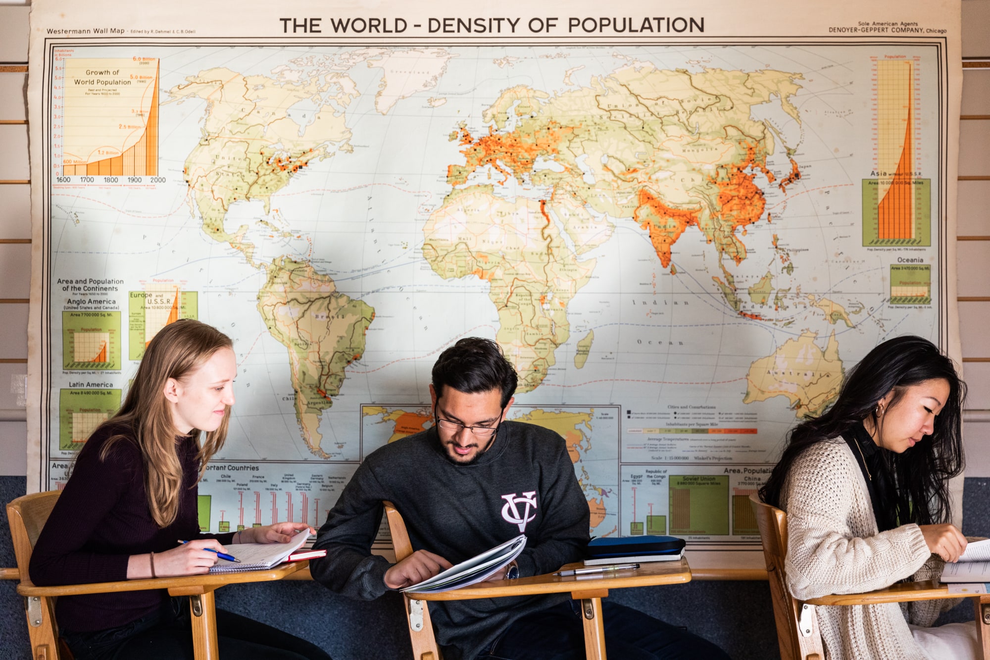 A student with dark hair and a mustache sits at a desk in a classroom, next to a giant map of the world.