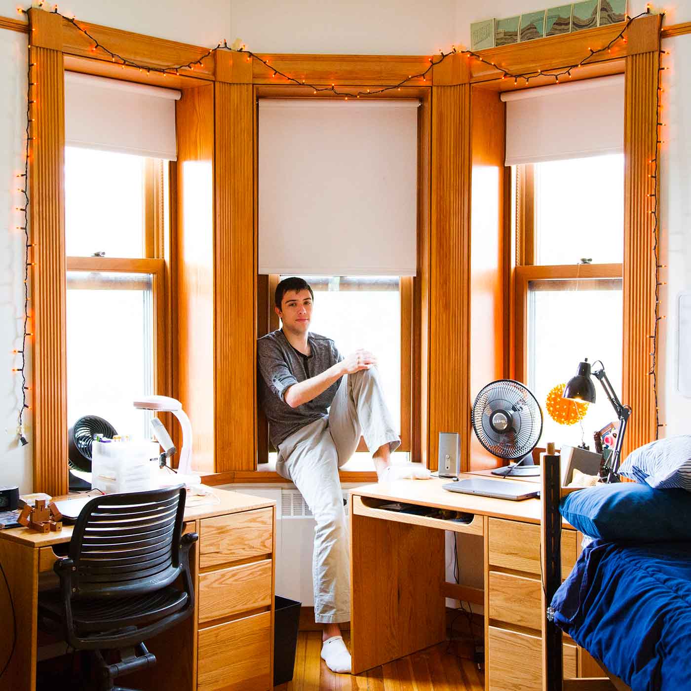 A student sits on a window sill between large, wood-framed windows, in a well-lit room. The student is looking at the camera.