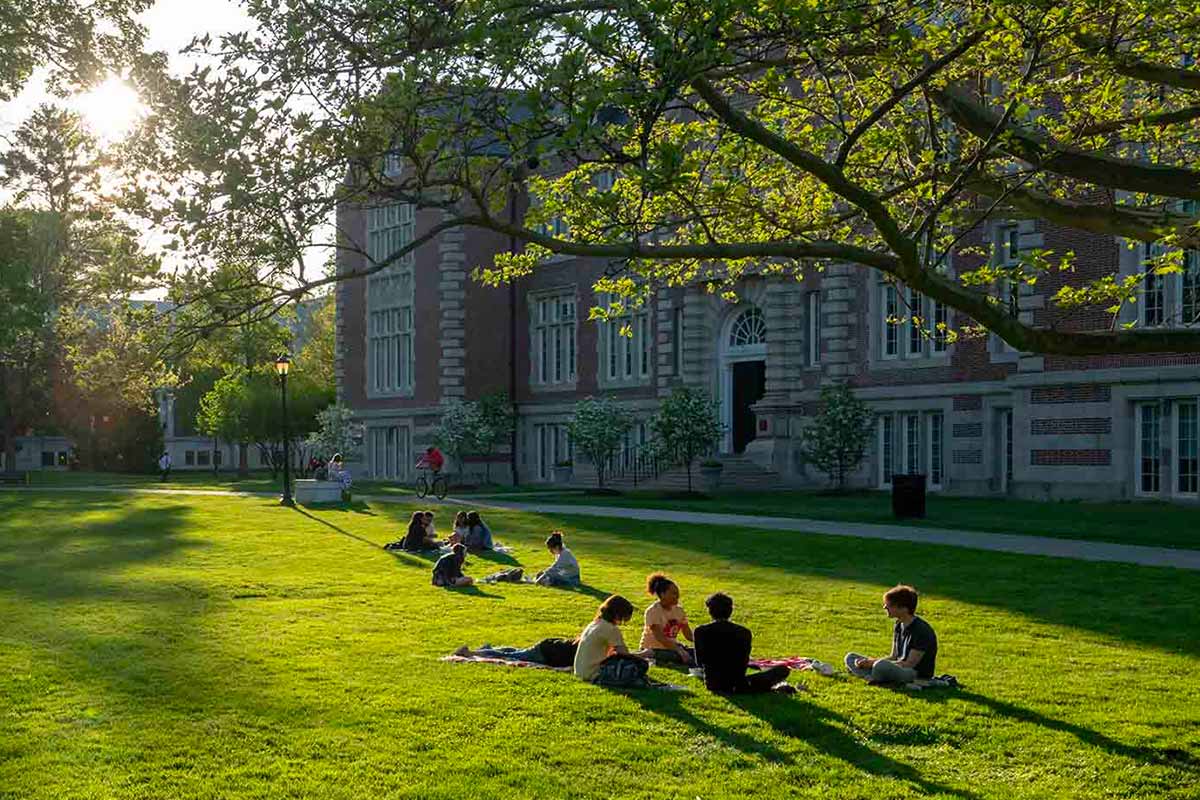 A group of people sitting on the grass in front of a large brick building in the late afternoon.