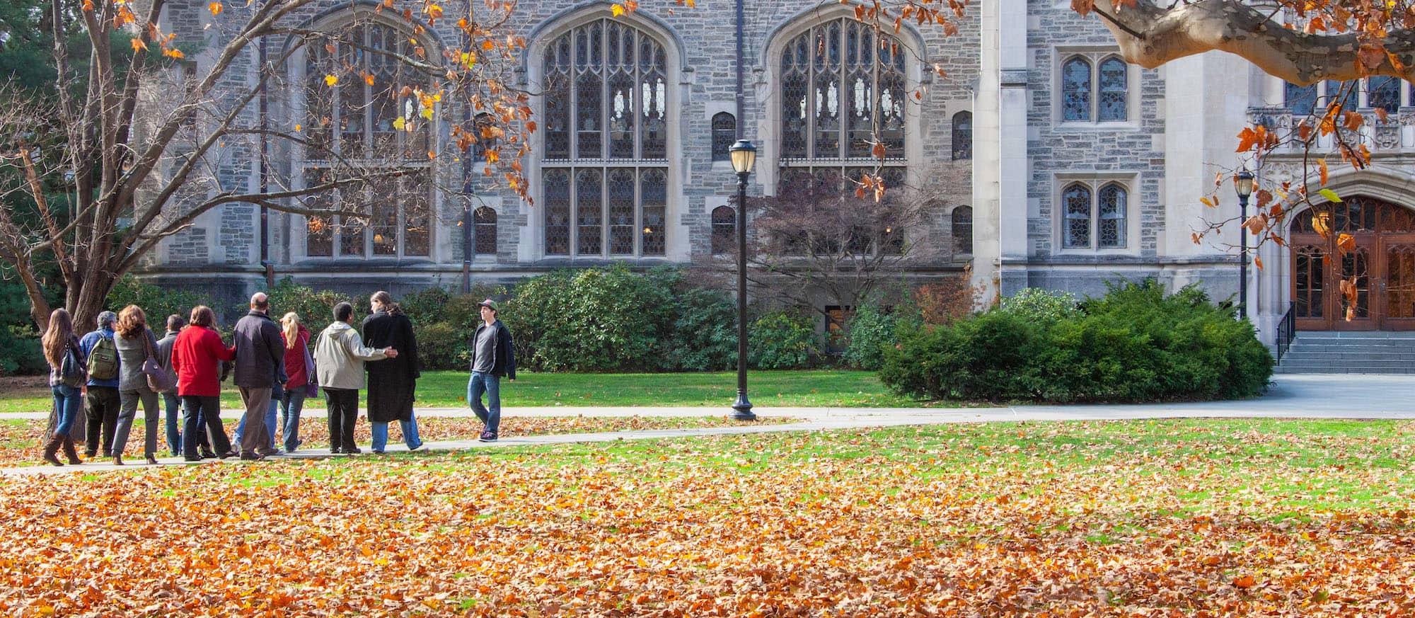 A group of people walk towards the Thompson Memorial Library, a stone building with large glass windows, on a fall day.