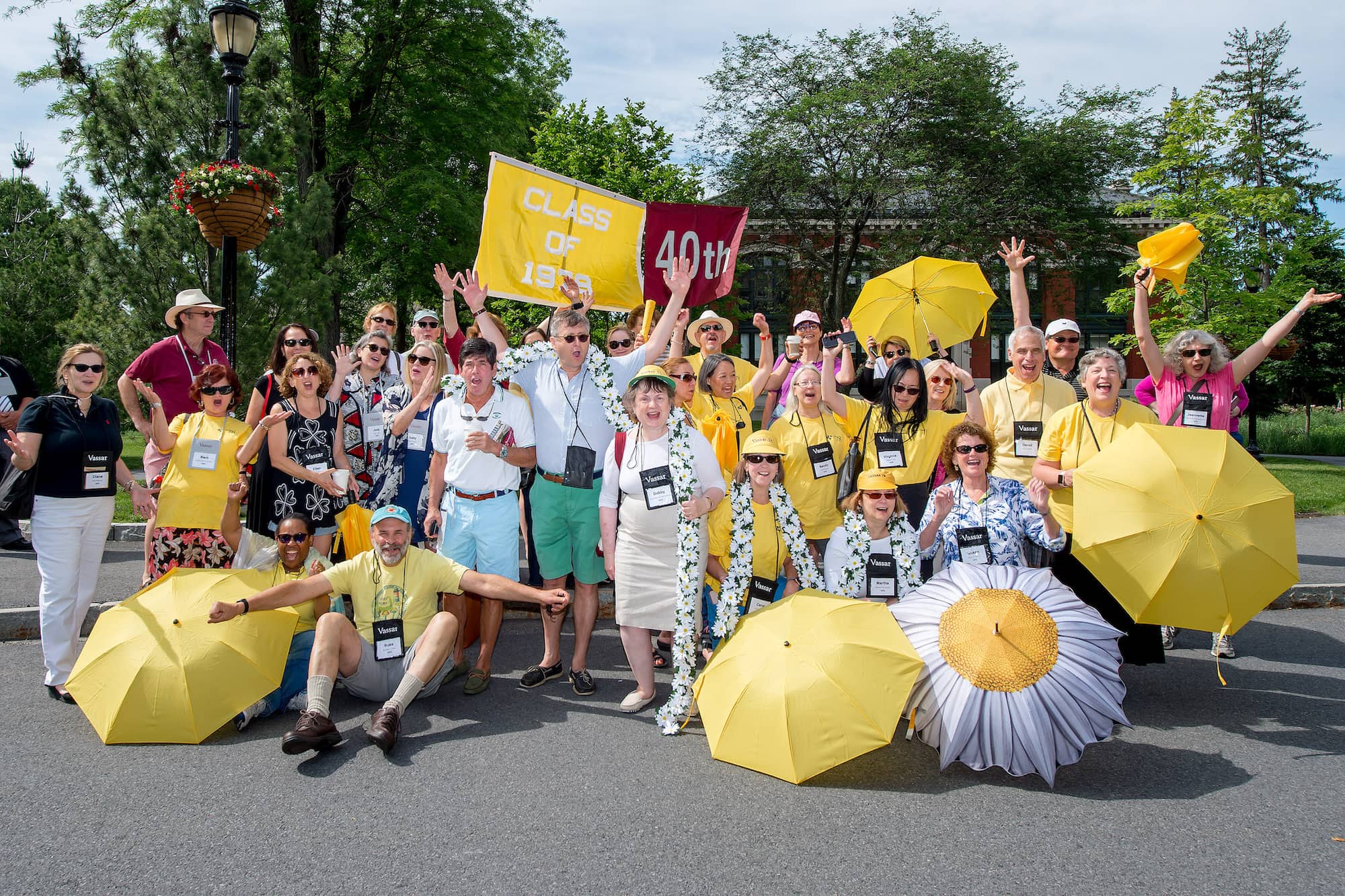 A large group of people are holding or sitting under bright yellow umbrellas or banners, wearing casual summer attire, cheering at the viewer.