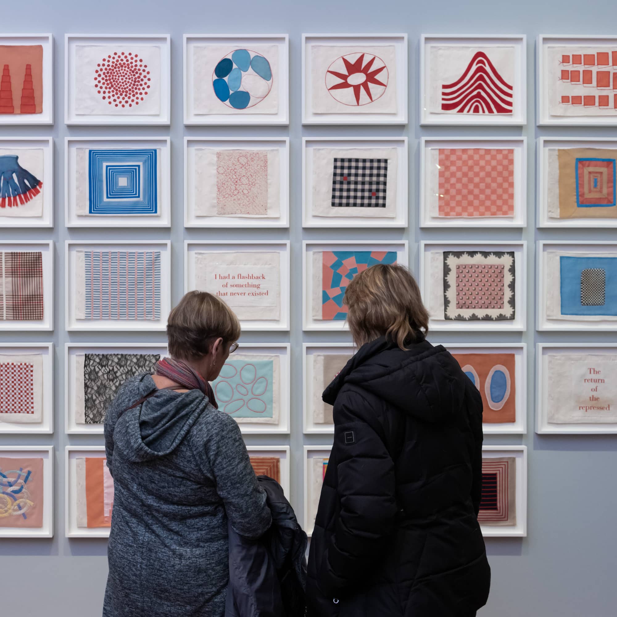 Two people, dressed in casual attire, are standing close to a wall, examining artwork consisting of multiple square frames, each containing different artistic patterns or designs, including abstract patterns, and varied color schemes.