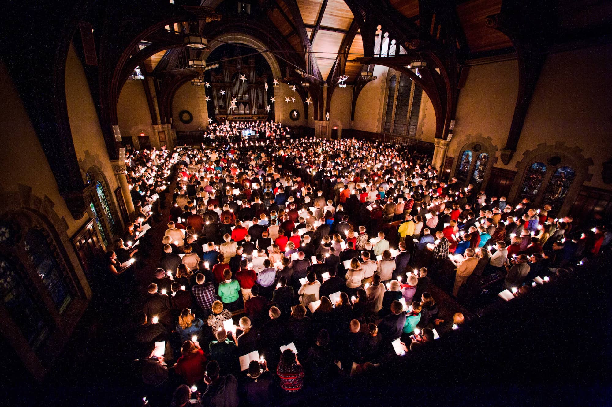 A large group of people seated inside a spacious church. Each person is holding a lit candle. The church’s gothic architecture is highlighted by the candlelight, revealing arched ceilings, wooden beams, and intricate designs.