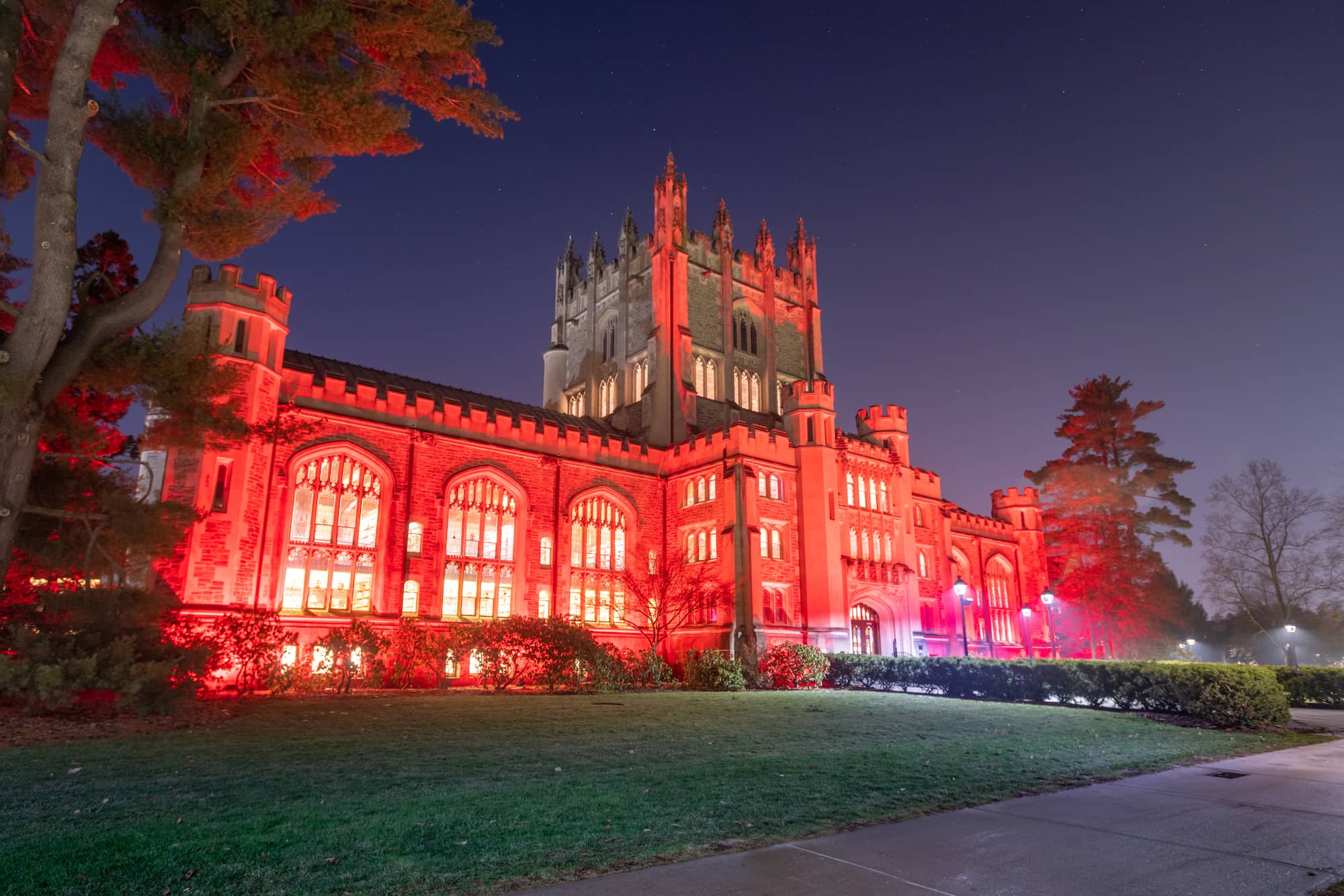 Vassar College Thompson Library, a large gothic-style building with arched windows, and detailed masonry, lit up in bright red at night, with lush green trees and well-manicured lawns in front.