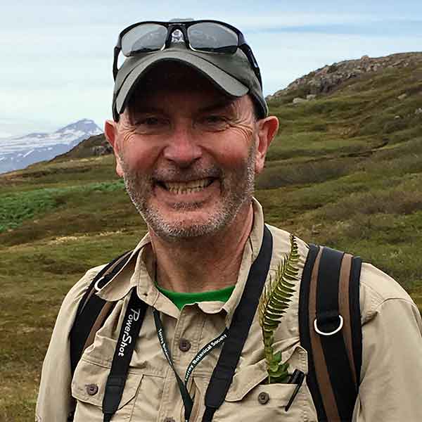 A person with a black baseball cap, a tan shirt, and a thin gray beard, smiles at the camera.