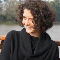 A smiling person with chin-length curly brown hair wearing a cowl-necked dark sweater.