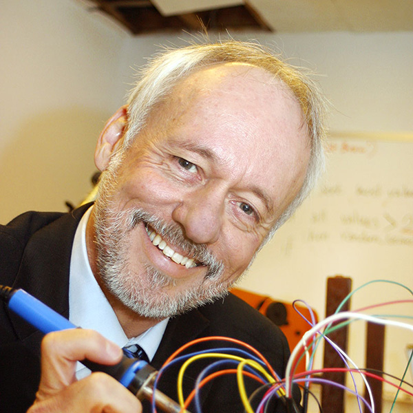 A person with short grey hair and a grey beard and mustache holding a soldering iron smiles at the viewer.