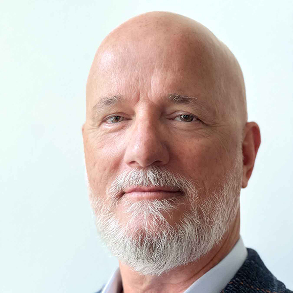 A person with a bald head and a gray beard and mustache, wearing a suit, looks at the viewer.