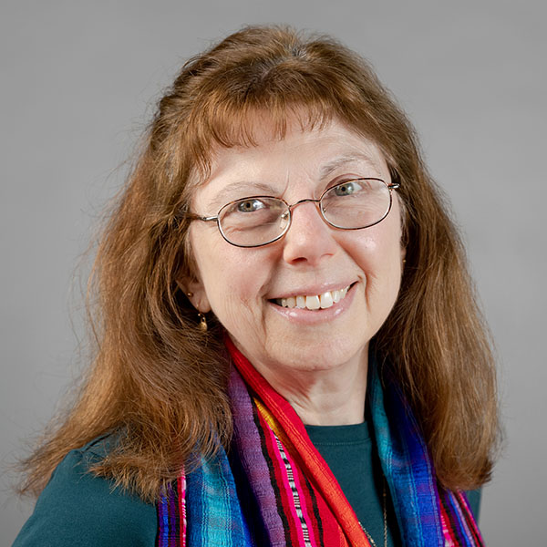 A person with long brown hair and bangs, wearing glasses, a teal sweater, and a multi-colored scarf, smiles at the viewer. Photo credit: Karl Rabe/Vassar College.